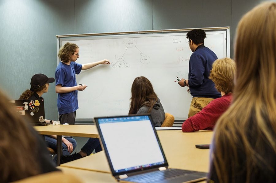 Students at laptop computers with students standing in front of a projection screen in a conference room gesture and confer together as they carry out an academic tutoring program in Michigan Tech’s computer science department.