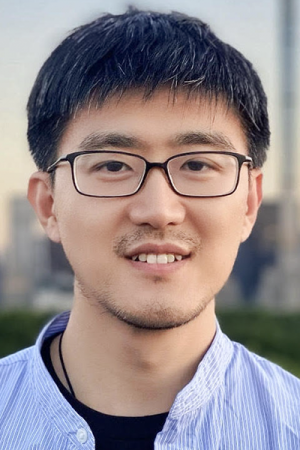 A man with glasses smiles at the camera with a blurred city background behind him.