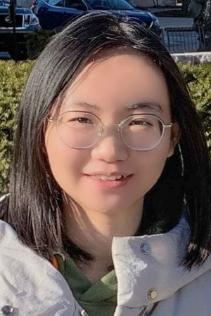 A woman wearing glasses smiles at the camera with a green seasonal streetscape, including parked cars, behind her.