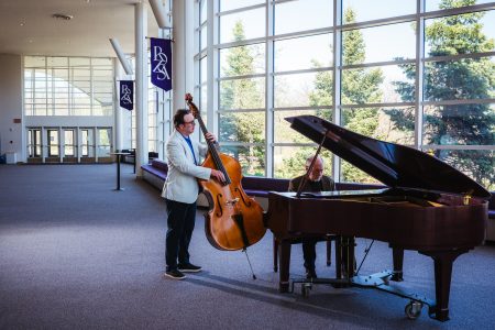 A standing bass player and a keyboard player seated at a grand piano play in the lobby of Michigan Tech’s performing arts center.