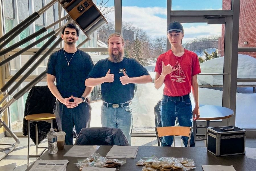 Three computing students stand in front of a window with a snowy campus outside giving a thumb’s up and a welcoming hand gesture at their bake sale on preview day at Michigan Tech.