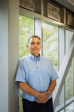 A Michigan Tech computing faculty member and institute director smiles in the walkway on campus between Rekhi Hall and the Library with a view of the outdoors.