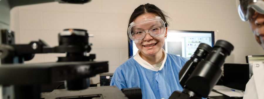 May Waters smiles at the camera, wearing safety goggles and a lab coat in front of a computer. A microscope and other equipment is in front of her.