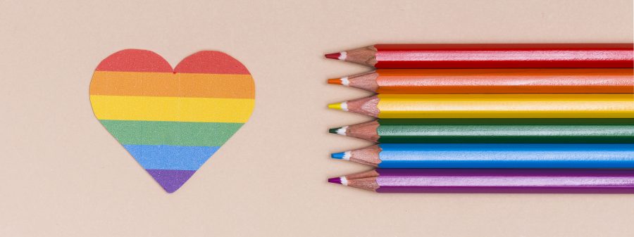A paper cut-out of a pride rainbow heart next to six colored pencils lined up in the pattern of a rainbow.