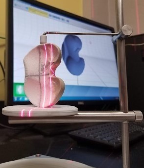 3D scan of tibial insert on a computer screen, with the actual tibial insert set in front of the screen in a vise, while being scanned