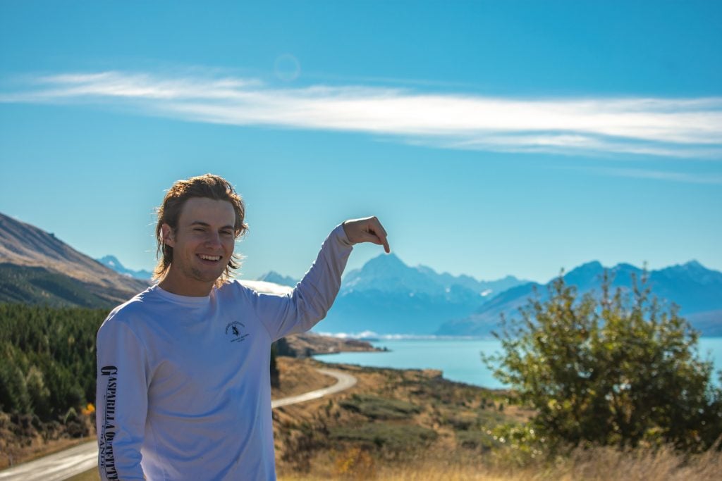 Ryan Schrader at the side of a winding mountain road, pointing at the mountain range in the background. His hand is curved and appears to touch the tip of the mountain.