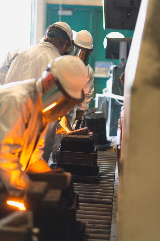 As the pouring team fills the 3rd mold [middle ground], an MSE staff member [foreground] lifts the mold jacket from the 2nd mold, and will transfer it to the waiting 4th mold [background] prior to it being poured. The jacket supports the green sand mold against the hydraulic pressure of the liquid metal entering the mold.