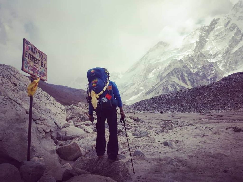 Sirak stands with backpack at Gorakshep, a small Himalayan Village at an elevation of about 16,942 ft. Note the iconic sign, "Way to Everest Base Camp".