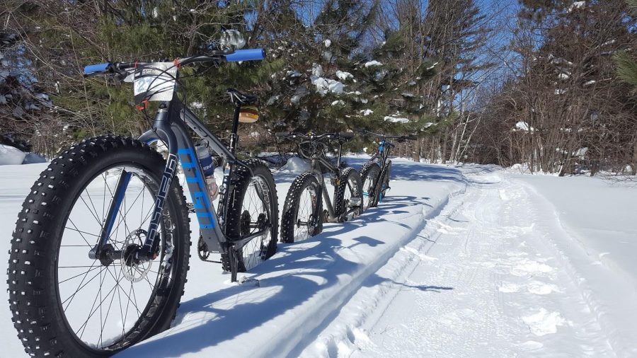 Three fat tired bikes are parked in the snow along the Michigan Tech "Tech Trails" groomed trail system, covered in snow, with sunshine and trees in the background.