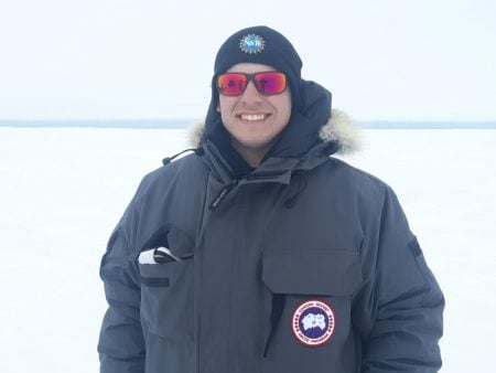 Caleb Kaminski in a parka with a knit hat and sunglasses outside in the snow.