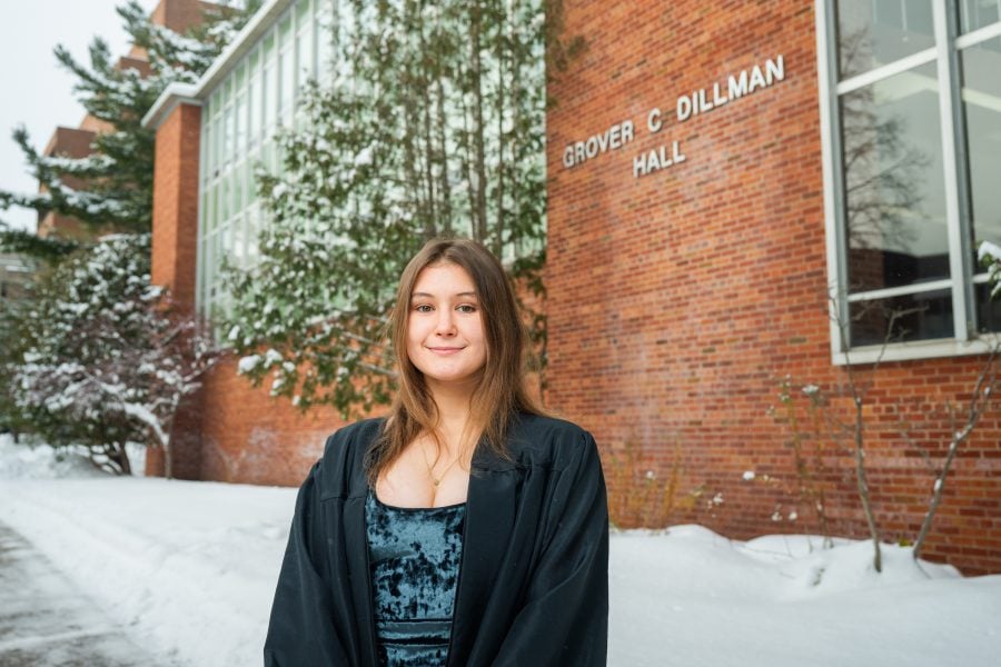 : Aliyah Maxwell-Abrams in their graduation gown standing in front of Dillman Hall
