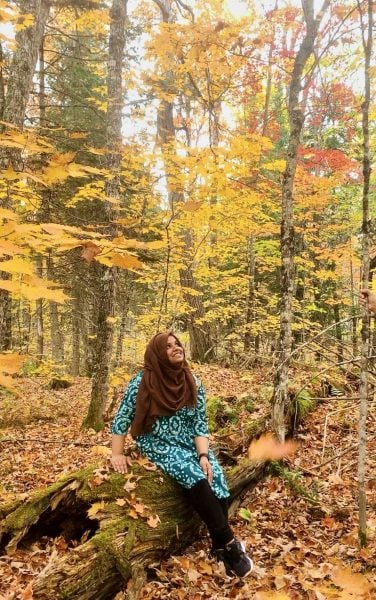 Mehnaz Tabassum seated on a log in the autumn woods surrounding campus