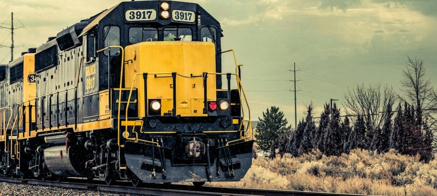 Front of a large black and yellow train driving down tracks with brush and trees in the background.