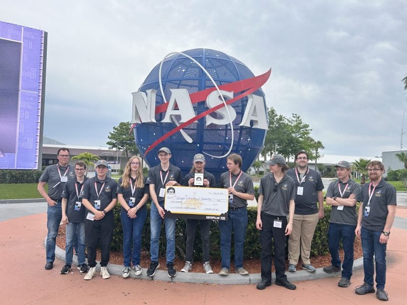 The MTU Astro-Huskies team poses in front of a NASA logo statue with their fourth place award at the Kennedy Space Center. The team is wearing gray Astro-Huskies polos and blue jeans.