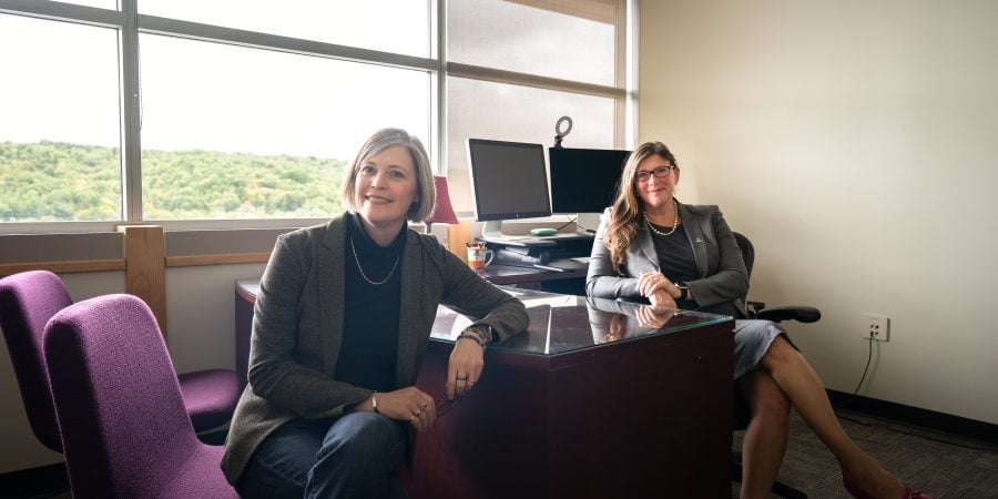From left, Audra Morse and Amy Landis pose at a desk in the College of Engineering Dean’s Office. Morse wears a charcoal blazer, black turtleneck sweater, and jeans and Landis wears a gray blazer and dress.