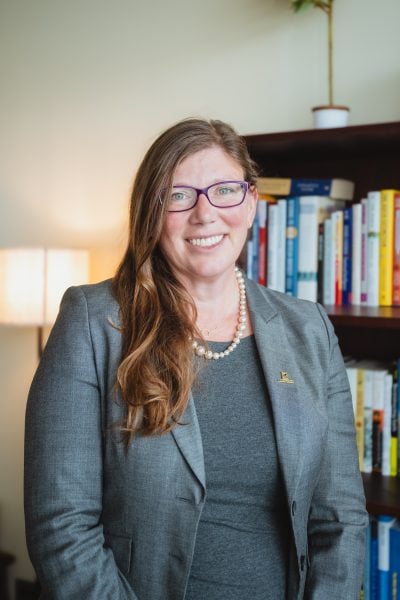Amy Landis smiles in a portrait wearing purple eyeglasses, a gray blazer with a Michigan Tech pin, and gray dress. 