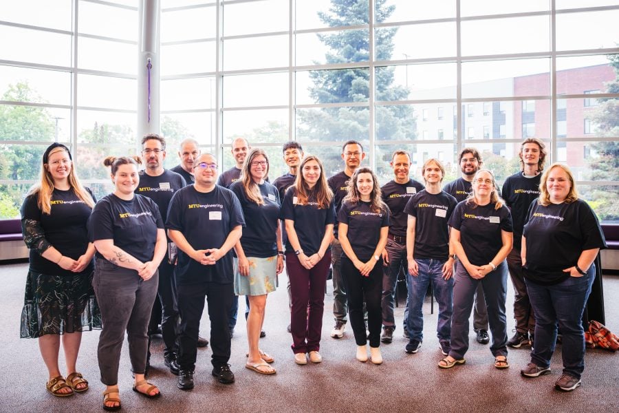 16 new faculty and staff gather for a group portrait in the lobby of the Rozsa Center for the Performing Arts. All wear the same black t-shirt that reads “MTUengineering” and stand in front of large windows.