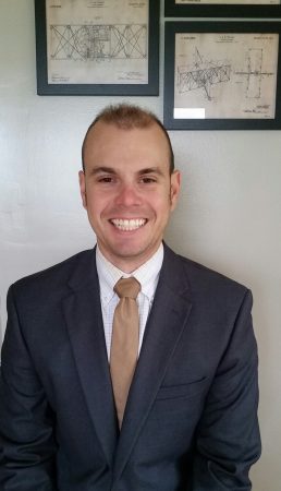 Kevin Howard smiles in a professional portrait wearing a blue suit, white patterned shirt, and tan tie.