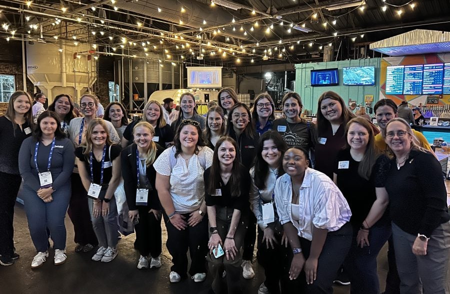 Dean Michelle Scherer and 22 Michigan Tech students and alumni pose together at an event as part of WE25. The group is wearing name tags and conference lanyards and business casual attire.