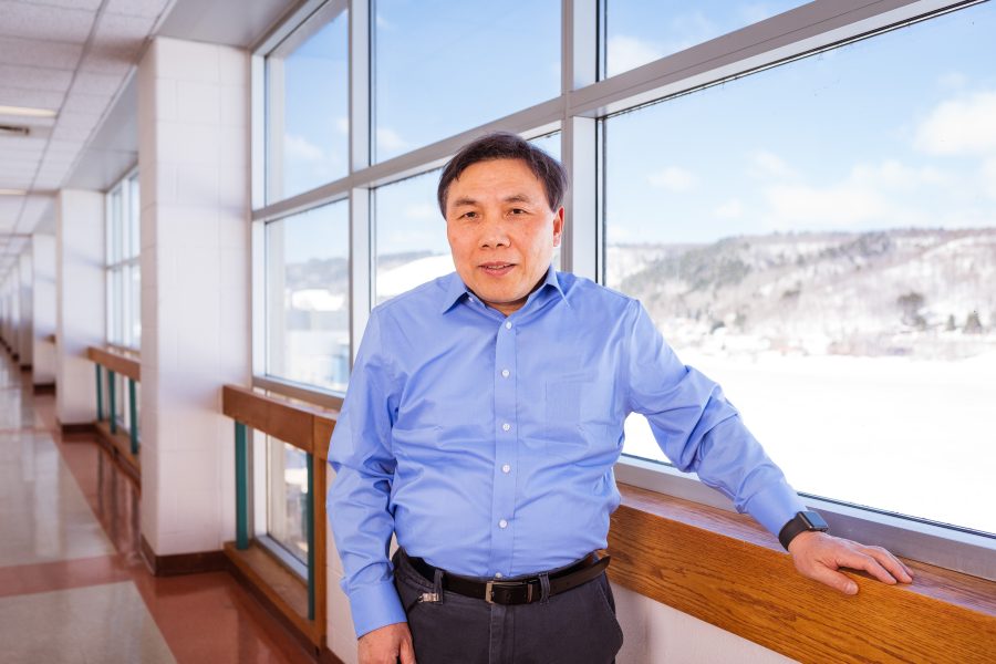 Yun Hang Hu stands in a hallway lined with windows showing snow-covered hills wearing a blue shirt.