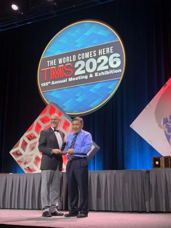 Two individuals stand on a stage holding a triangular glass trophy under a sign that reads “The World Comes Here, TMS 2026, Annual Meeting and Exhibition.”