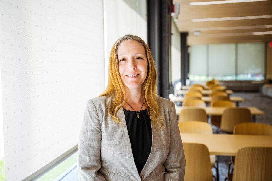 Portrait of Adrienne Minerick wearing a black shirt and grey blazer in front of an academic lobby with tables and chairs. 