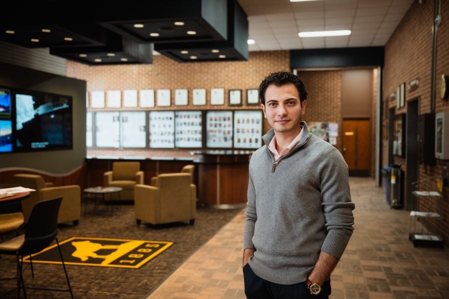 Man in a gray sweater stands confidently in a modern office lobby with brick walls and comfortable seating areas.