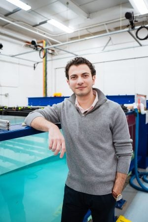 Man in a gray sweater leaning on a large blue industrial tank inside a brightly lit workshop.