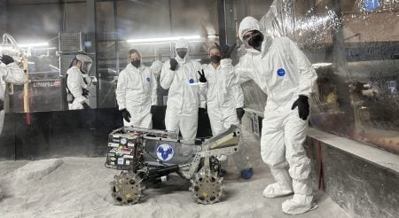 Michigan Tech students pose in protective equipment with their lunar rover prototype in a simulated lunar environment.