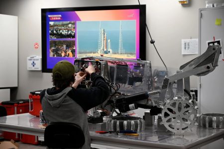 Michigan Tech student works on lunar rover in lab space with the Artemis II launch stream playing on a mounted TV set.