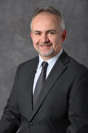 A professional headshot of M. Emin Kutay wearing a charcoal suit in front of a gray backdrop.