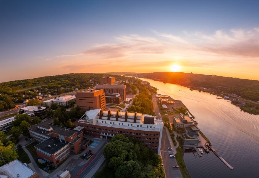 Aerial view of Michigan Technological University at sunset. The campus buildings are surrounded by lush greenery, and a wide river is visible on the right side, reflecting the sun's golden light.