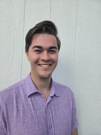 Headshot of Connor MacRobbie wearing a purple button up shirt in front of a white wood background.