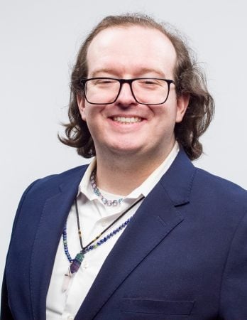 Headshot of Duncan Mullins wearing a blue blazer, white shirt, and beaded necklaces in front of white background.