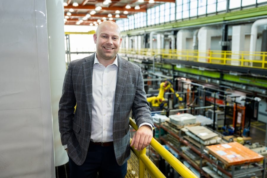 Joseph Williams stands on a walkway above a manufacturing plant floor, wearing a plaid gray blazer and white button down shirt.