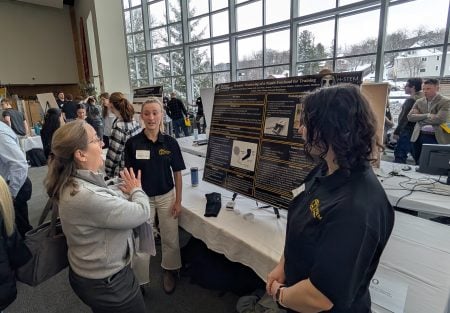 three people standing in front of a table with a project poster displayed and people walking around behind them