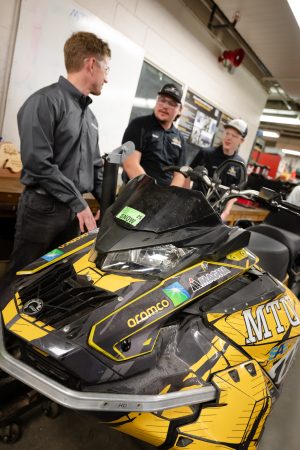 Aramco's Andrew Baur talks to two Michigan Tech students standing next to a snowmobile in the Machine Shop