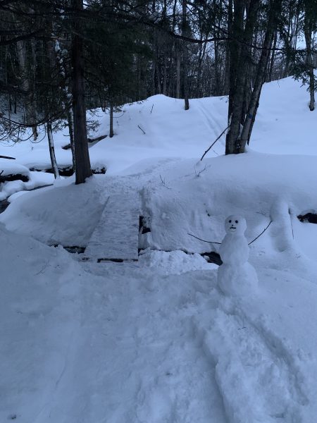 A snowman in front of a wood plank bridge over a stream in a snowy river