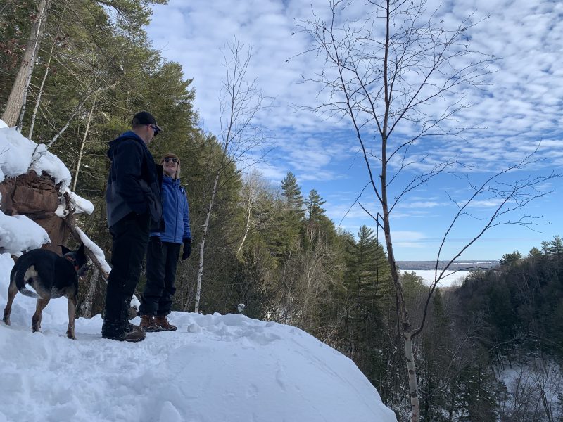 Two people looking at each other with a dog on a cliff surrounded by forests