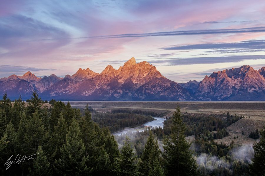 A mountain at sunset overlooking fields and a coniferous forest with a river winding through it