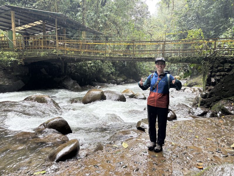 Person standing next to waterfall giving a thumbs up gesture. 