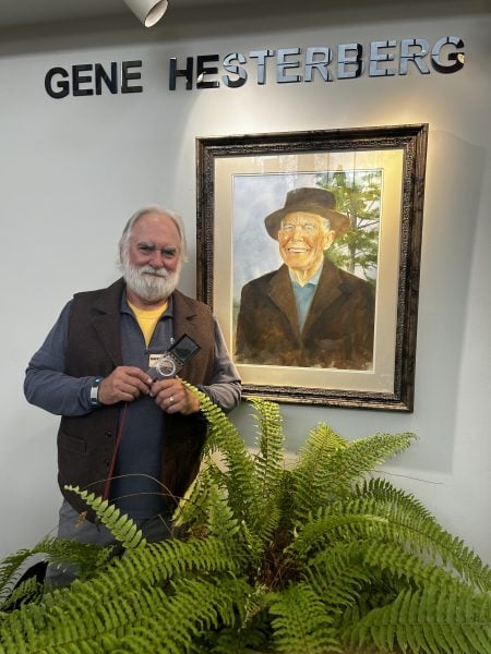 RJ Laverne holding a compass stands next to a painting of Gene Hesterberg.