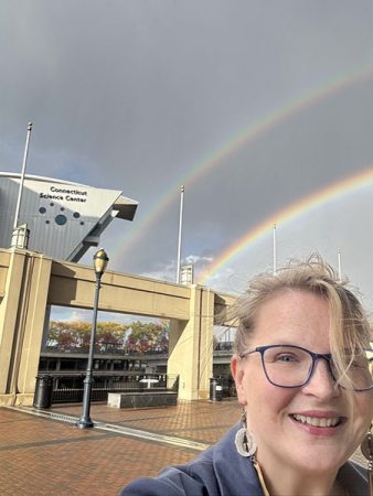 Tara Bal poses outside of the Connecticut Science Center, a double rainbow framed in the sky above the Center.