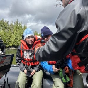 Quinn Riordan and her stepmom sit on the edge of a raft wearing life jackets and outdoor gear. They are leaning towards the camera to get a better view of the red rock crab held by their guide.
