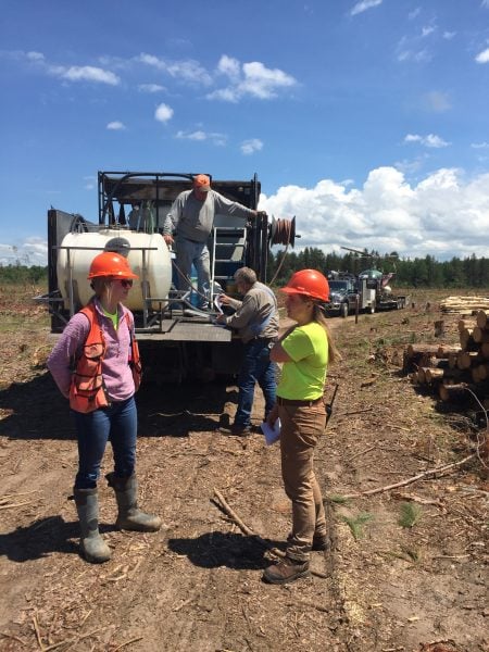 Amber Marchel stands in the foreground speaking with Claire Runquist on a dirt road through harvested timber woodland. Behind them, two site contractors work in the bed of a truck equipped with a large barrel and hose.
