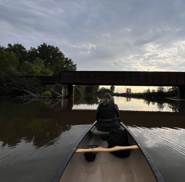 A point-of-view shot from the back of a canoe shows a student sitting at the front, facing away from the camera towards the sun rising over the Sturgeon River. The canoe is headed down the river towards a bridge. The river is lined on either side with trees and brush.