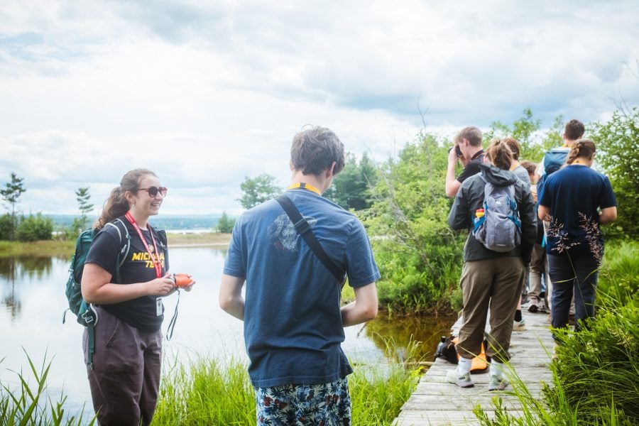 Sierra Williams stands outdoors wearing a Michigan Tech t-shirt, smiling as she guides SYP students along a boardwalk wetland trail.