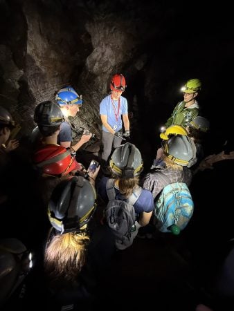 Bre Gusick stands in a cave surrounded by students. All are wearing hard hats and head lamps. Gusick carefully holds a bat for the students to see.