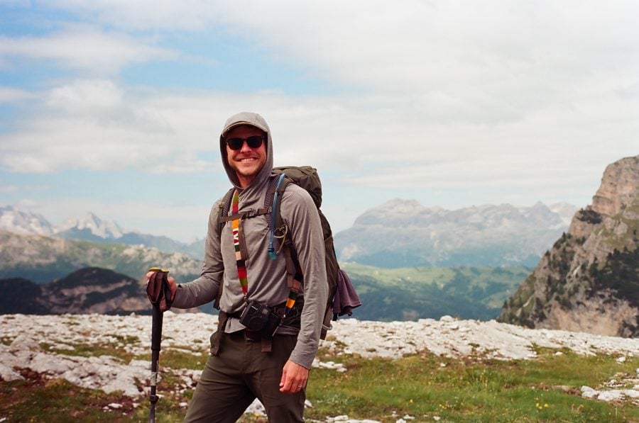 Christian L. John smiles at the camera, wearing sunglasses and hiking gear as he treks across a mountainous landscape dotted with stones and wild grasses.