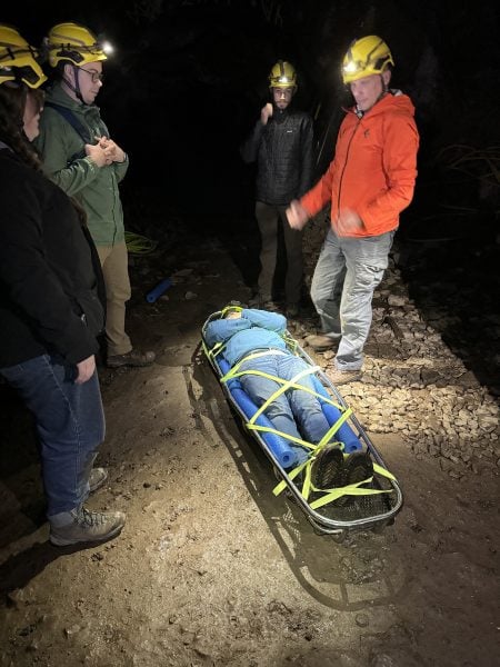 Four men stand in a mine underground, demonstrating how to safely secure a person a a stretcher in the dark with only headlights illuminating.